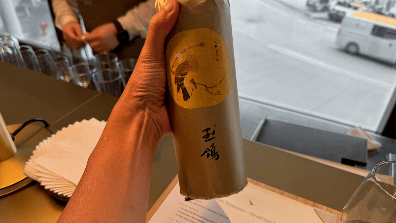 一隻手持著標有「玉鴿」字樣及鳥類圖案的酒瓶,背景為餐廳環境。