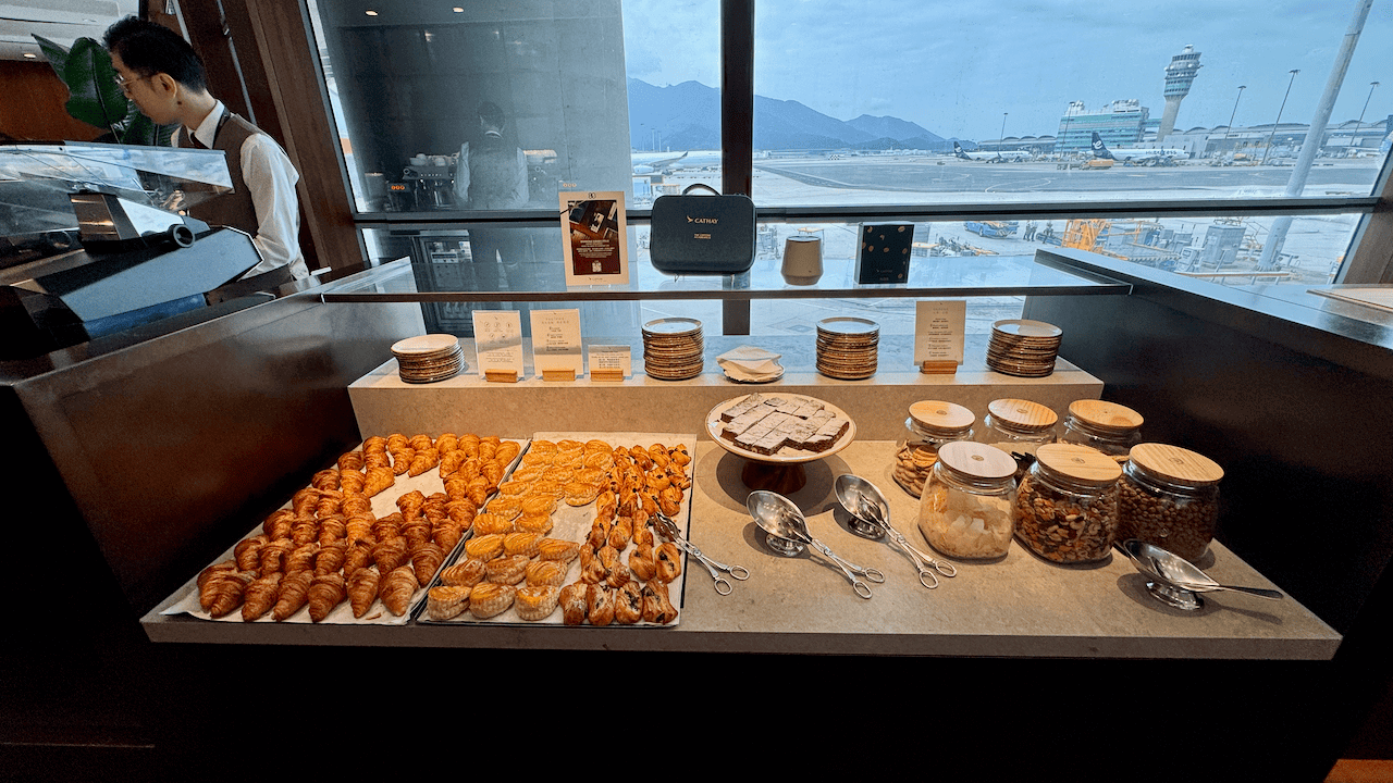 國泰航空貴賓室自助餐檯,擺放牛角包、糕點及堅果,窗外可見機場停機坪景色